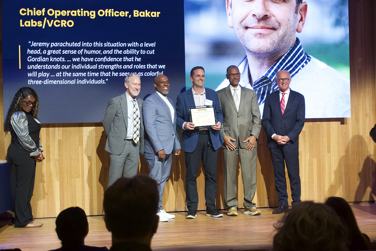 Five men stand on stage posing for a photo -- the one in the center holding a ceremonial certificate. A woman, assisting with the event, stands off the side to the left.