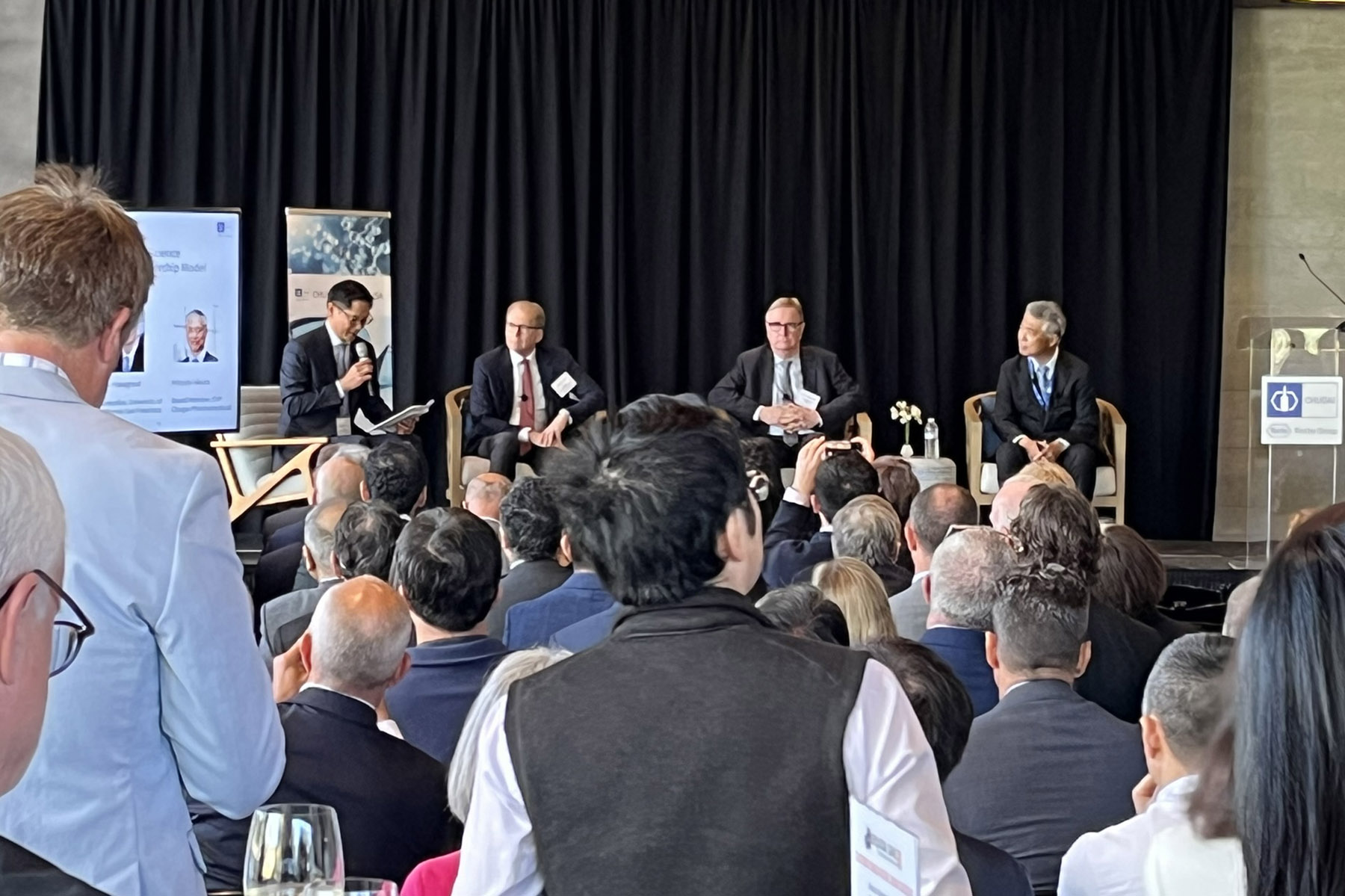 Four men in a panel presentation sit on stage in an auditorium filled with people