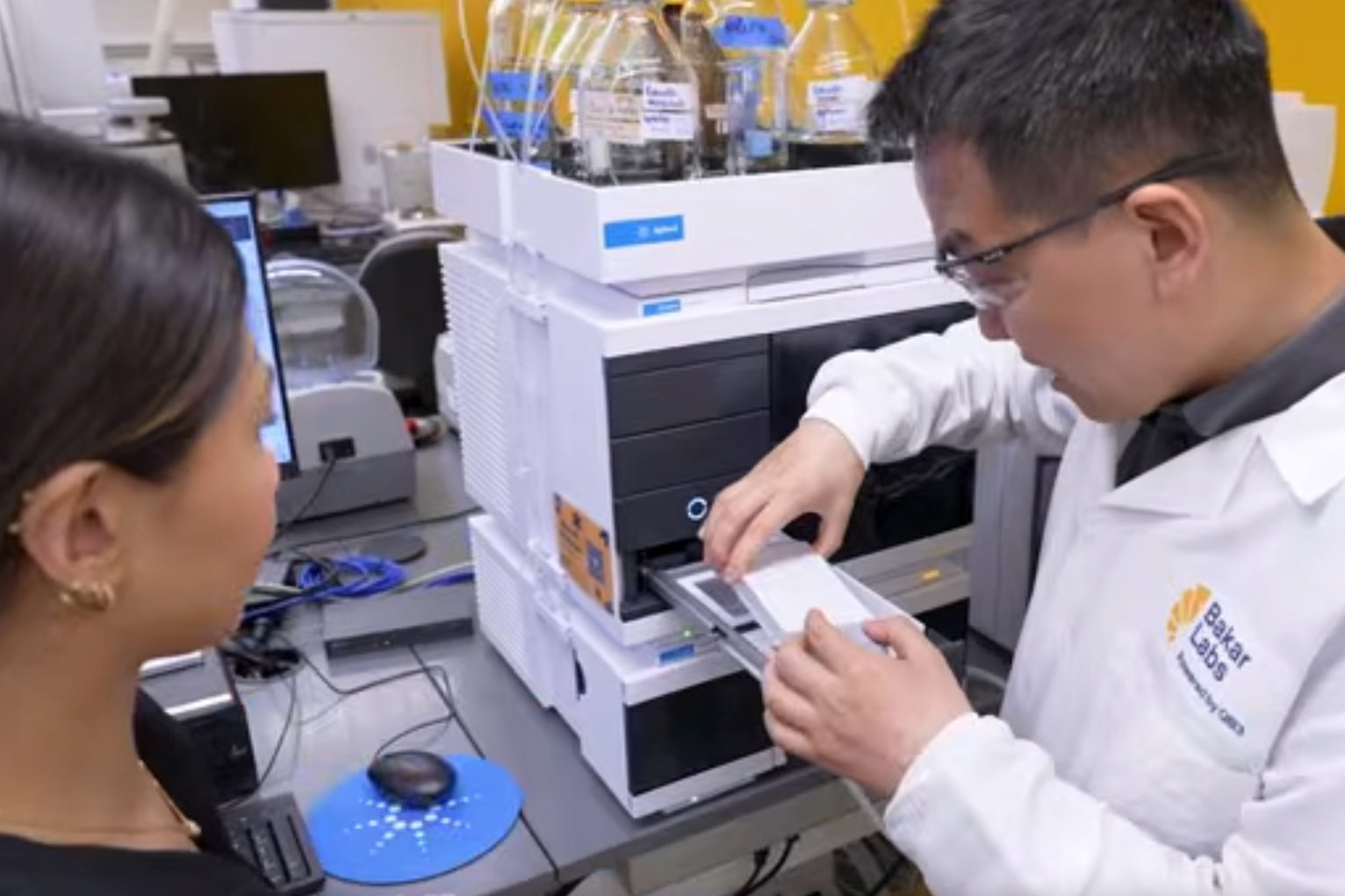 Woman in black watches man in lab coat load sample into equipment